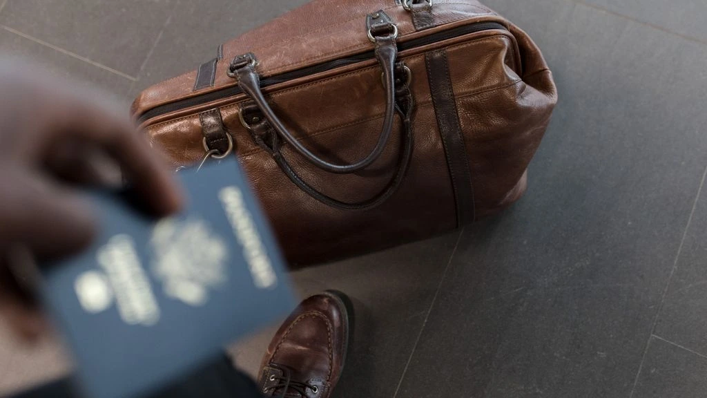 Leather carry-on suitcase, boarding pass, and tortoiseshell sunglasses on cream textile